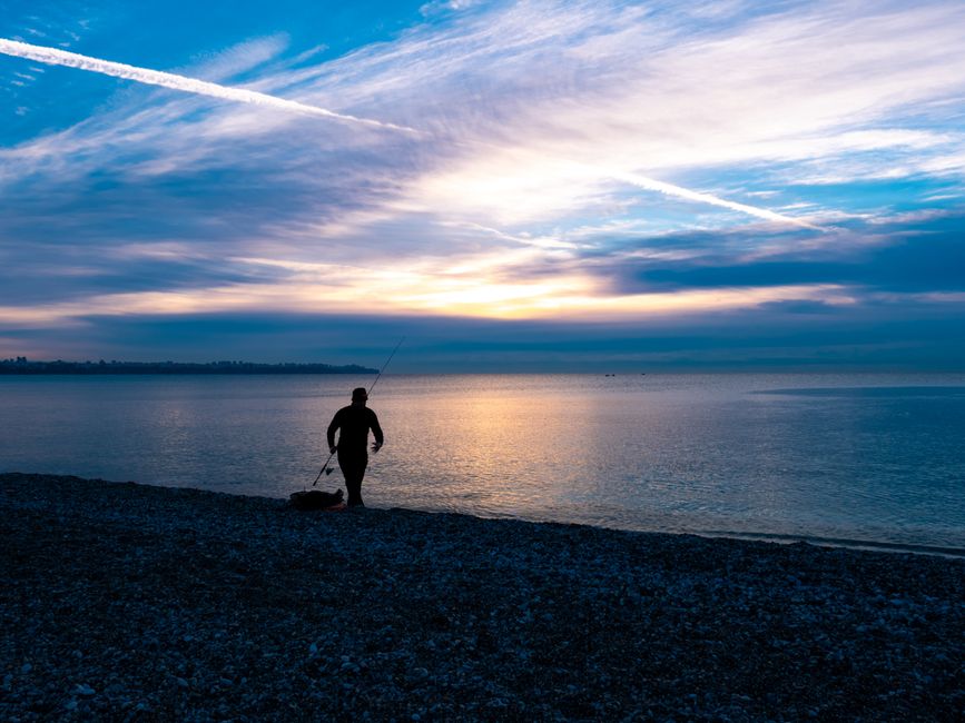 Junger Angler wirft eine Spinnrute vom Seeufer bei Sonnenaufgang, weite Landschaft mit ruhigem Wasser, mit einer Angelrute für Anfänger.
